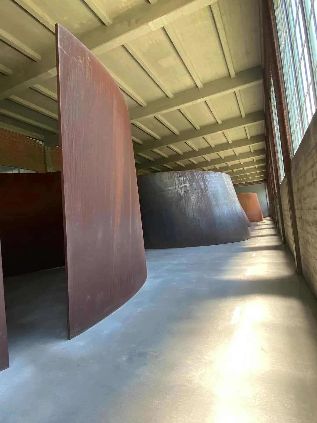 Interior gallery space with polished concrete floor and large burgundy and dark metal curved sculptures in an industrial warehouse setting.