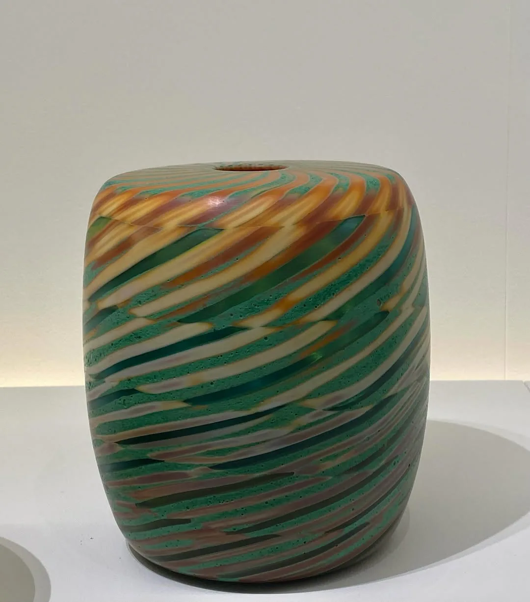 Ceramic vessel with diagonal striped pattern in green, gold, and terracotta tones, cylindrical form with textured surface.