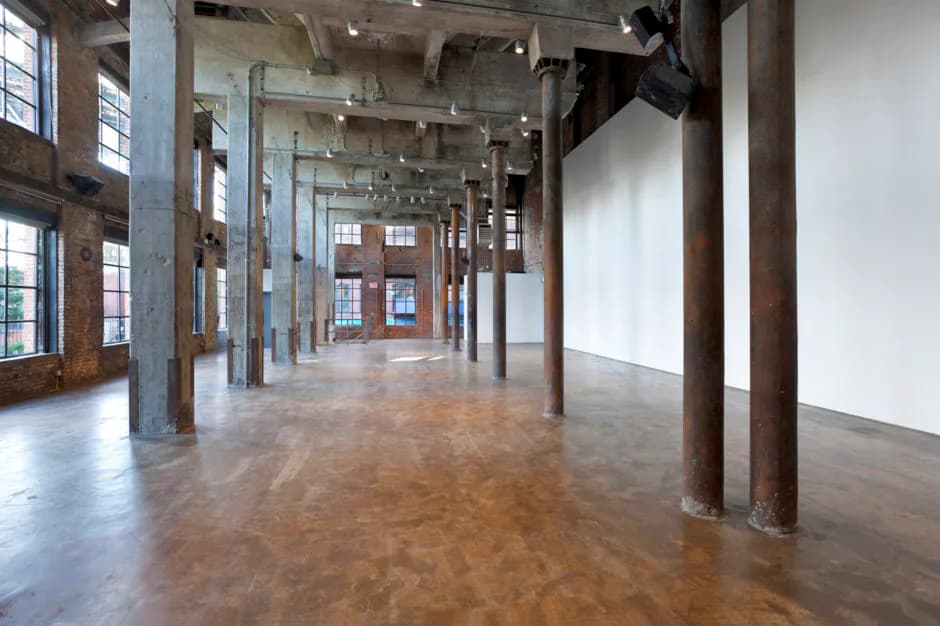 Industrial loft gallery interior with polished concrete floors, exposed brick columns, large windows, and white walls.