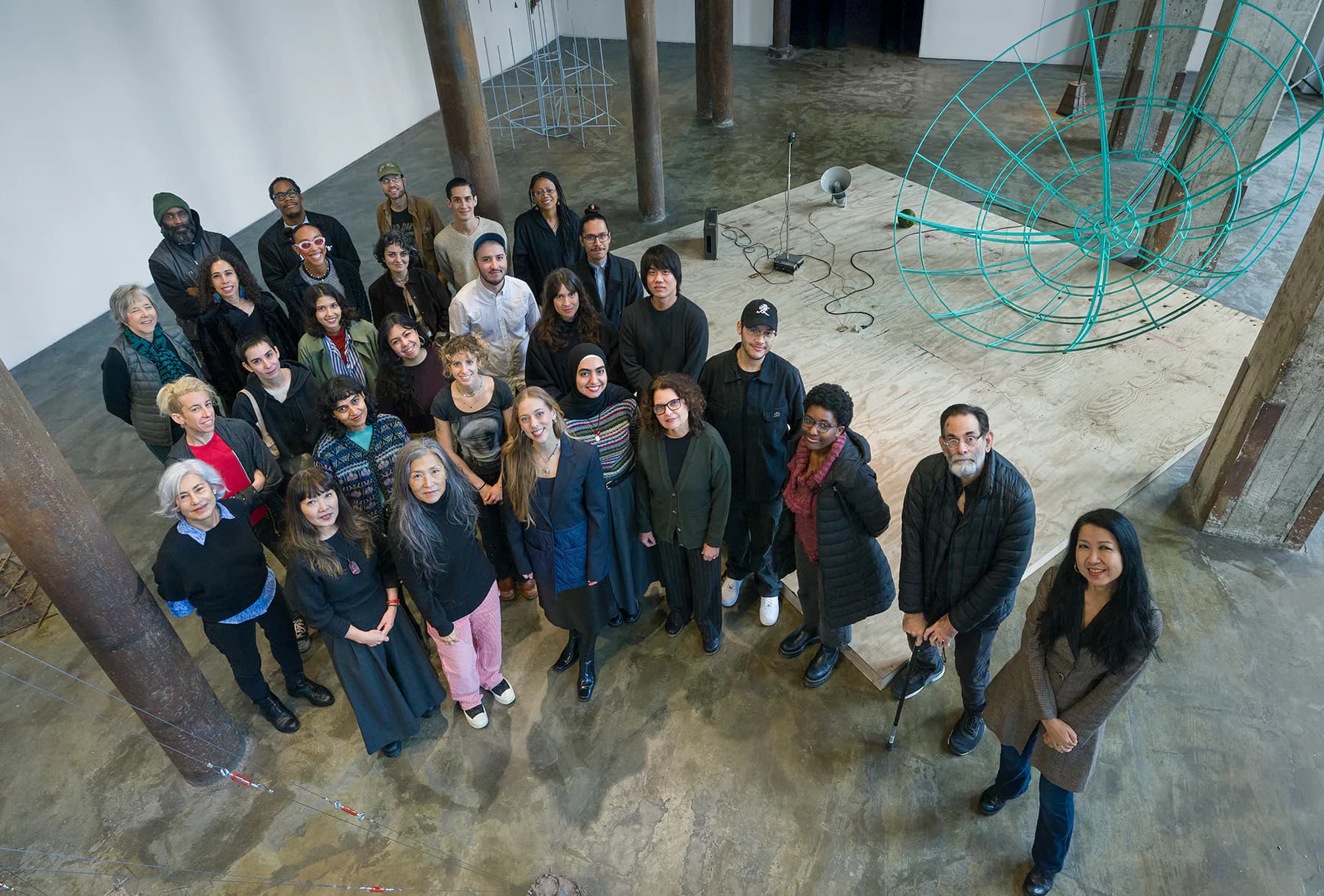 Group of approximately 30 people standing in an unfinished industrial gallery space with concrete floors and green sculptural elements.