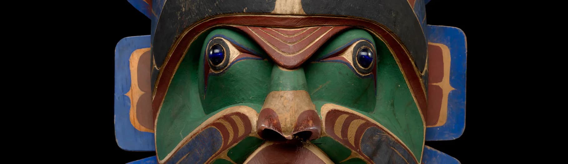 Carved wooden mask with green face, prominent mustache, and copper-toned accents, displayed against blue and orange background.