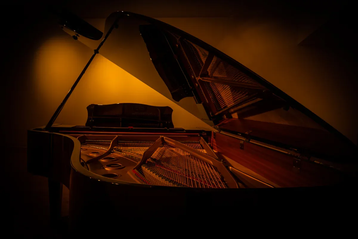 Grand piano with its lid propped open, lit dramatically from above against a warm golden-brown background, emphasizing the instrument's interior strings and wooden construction.