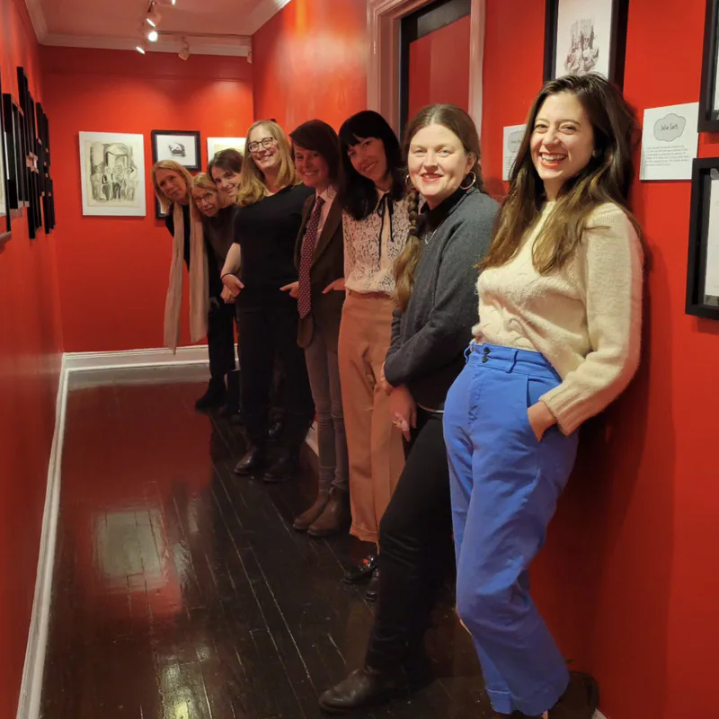 Group of people standing in a gallery with bright red walls displaying black-and-white framed artwork and prints.