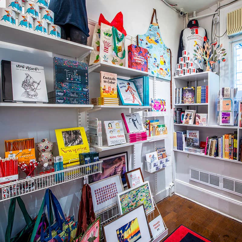 White shelving unit displaying illustrated children's books, prints, art prints, and colorful merchandise in a bright gallery or shop interior.