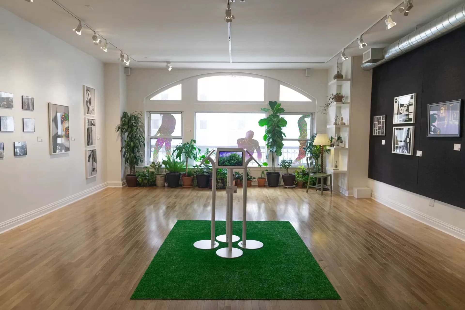SoMad gallery interior with wooden floors, white walls, black accent wall, large windows, track lighting, potted plants, and green artificial turf installation in center.