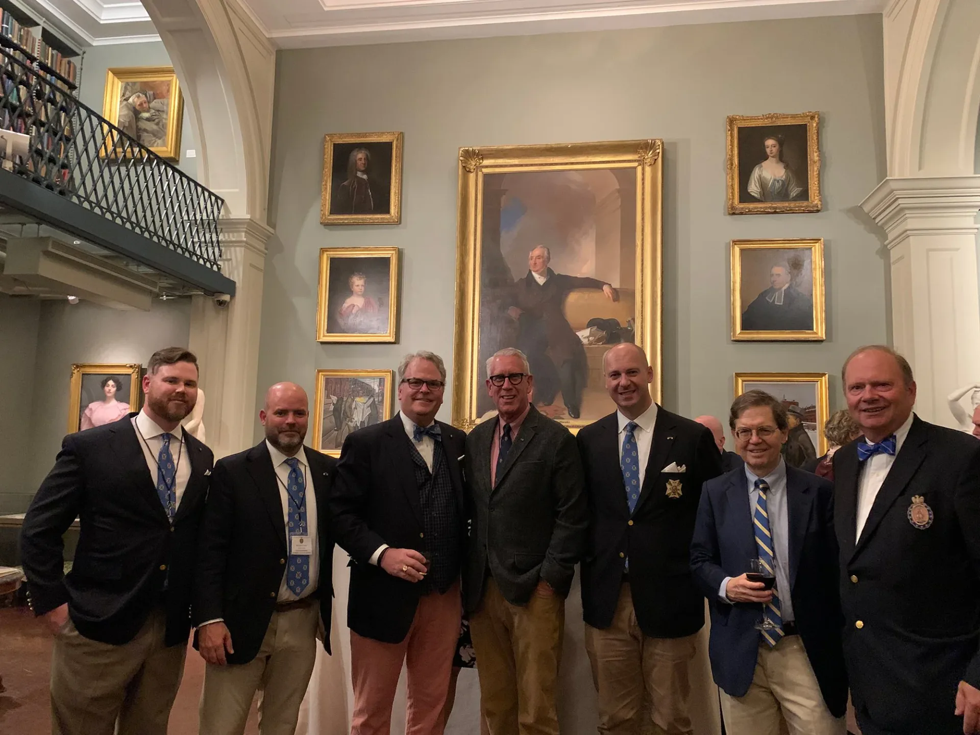Seven men in formal attire stand in an elegant gallery interior with gilt-framed portraits on beige walls and white columns.
