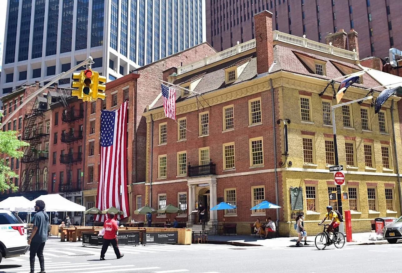 Historic brick and yellow building with multiple stories and American flags, surrounded by modern skyscrapers in an urban plaza.