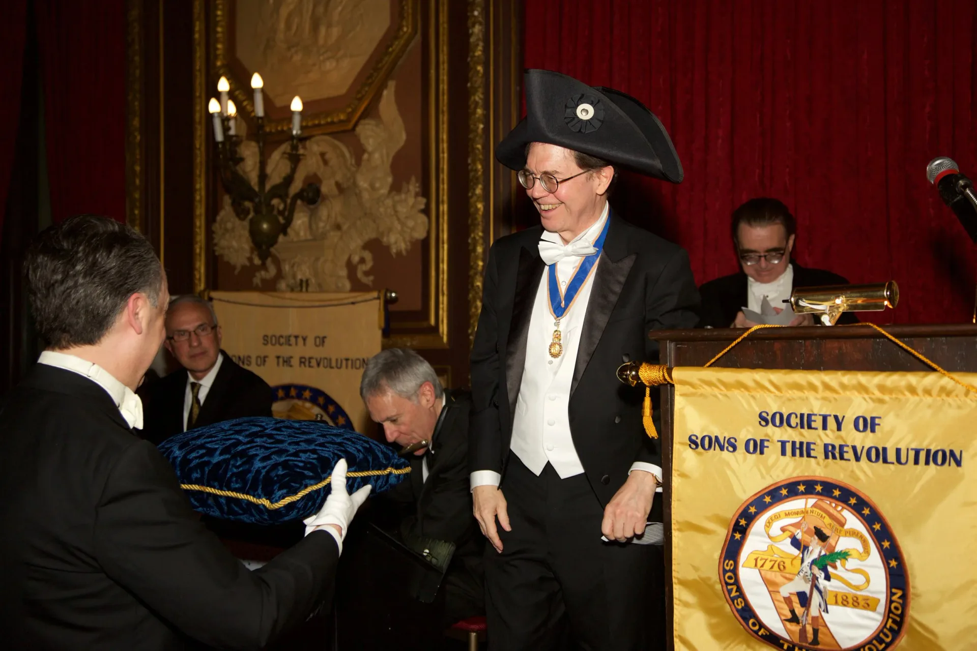 Man in formal attire and tricorn hat standing at podium marked 'Sons of the Revolution' in ornate red-walled interior with candelabra.