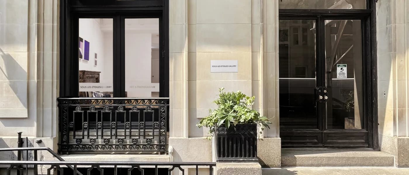 Storefront of Sous Les Etoiles Gallery in Manhattan with black-framed windows, cream-colored facade, and a potted plant by the entrance.