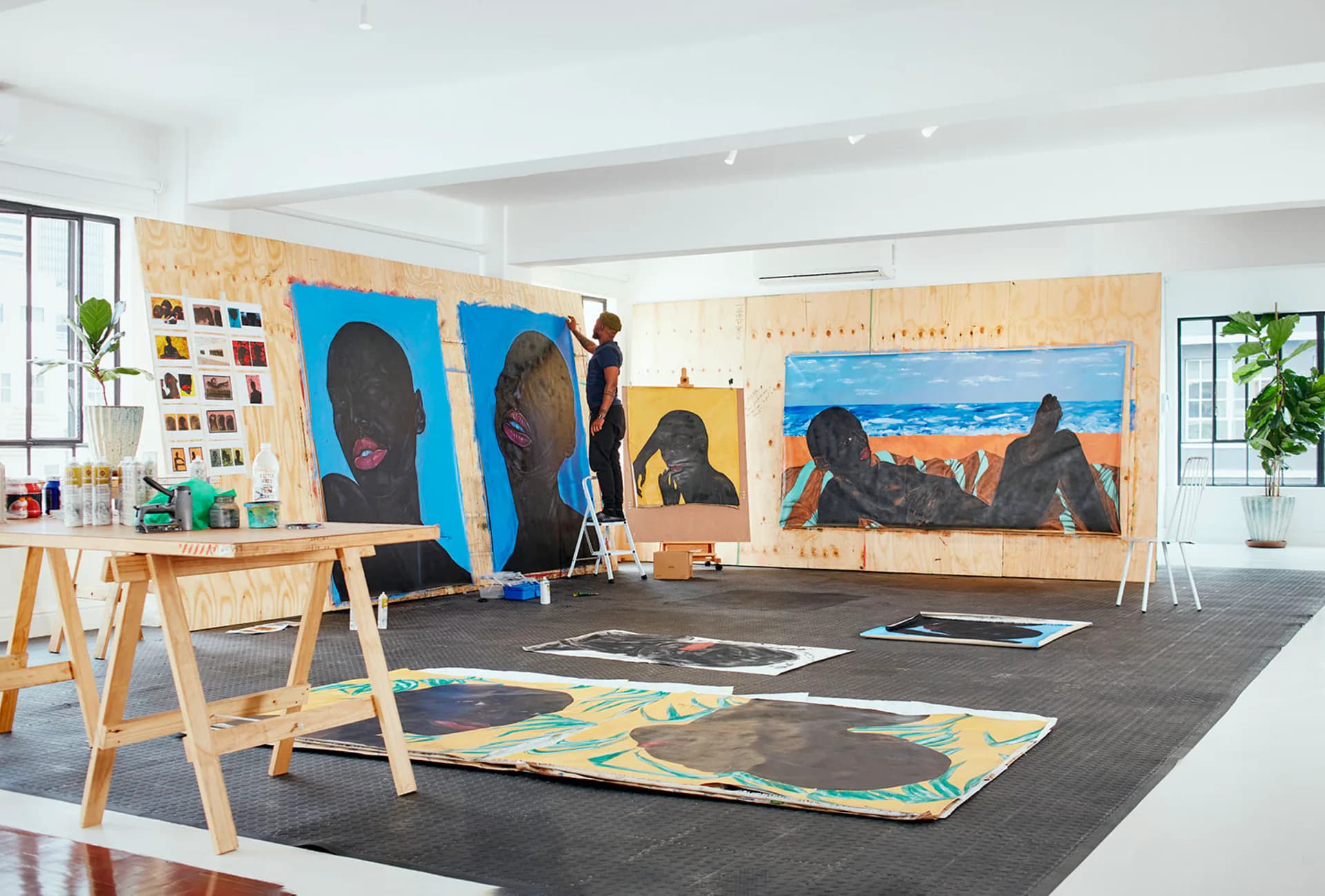 Southern Guild gallery interior with large blue portrait paintings on wooden panels, wooden work tables, and colorful artworks displayed on dark flooring.