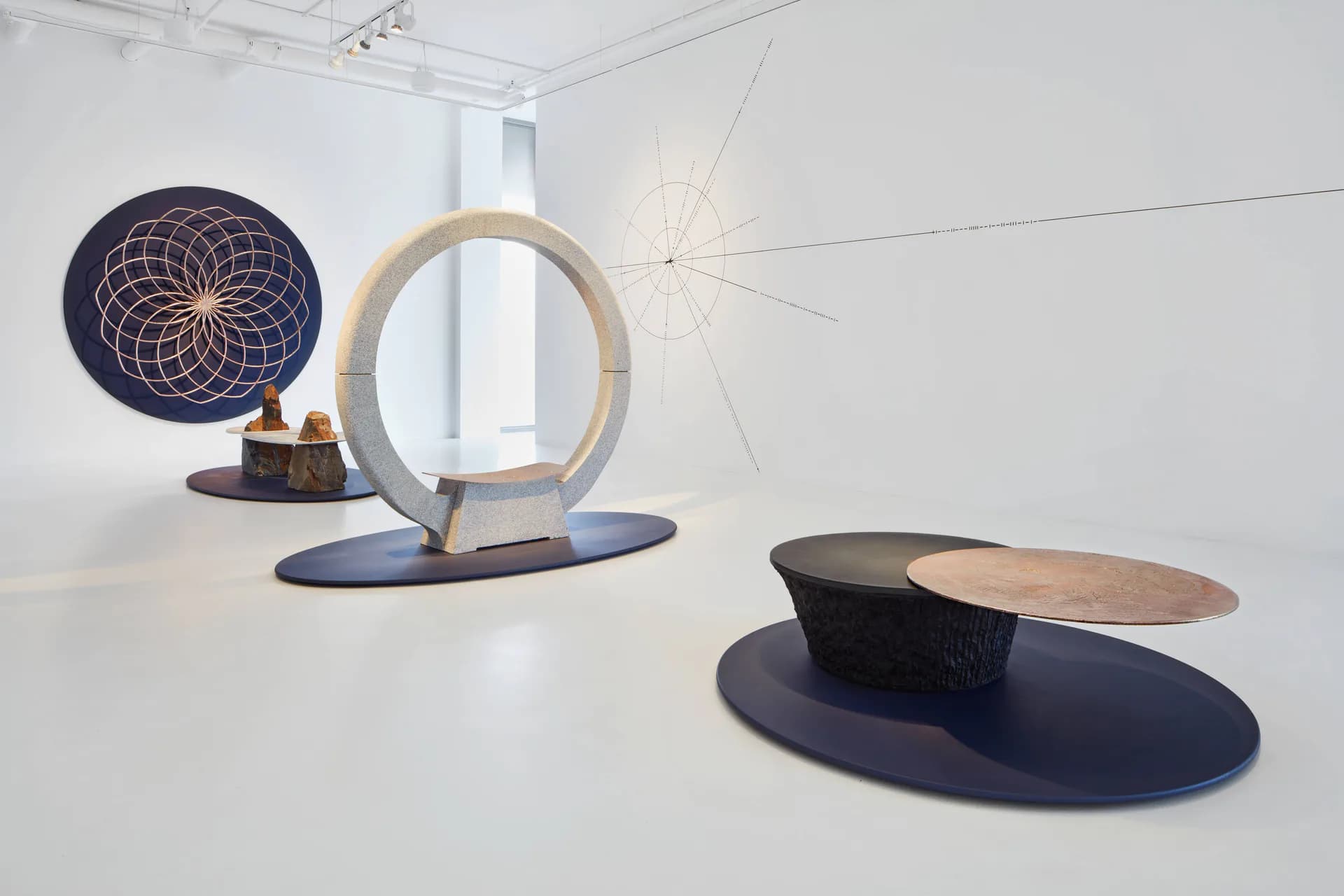 Abstract sculptural installation featuring wooden and metal circular forms, geometric shapes, and navy blue curved elements arranged on white gallery floor.