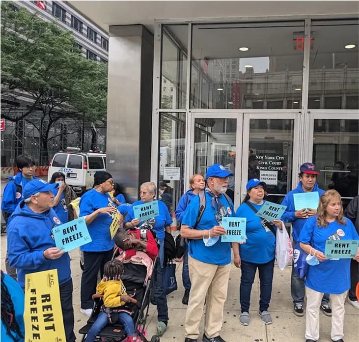 Exterior photograph of an urban storefront with people in blue shirts holding signs during what appears to be a community event or protest.