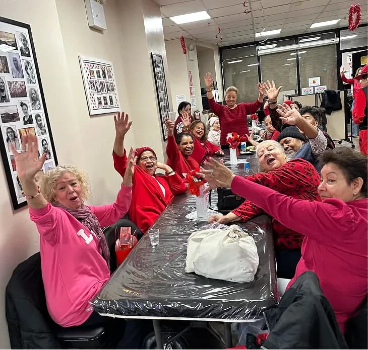 Interior photograph of people in red and pink clothing gathered around a table in a community space with photographs on the walls.