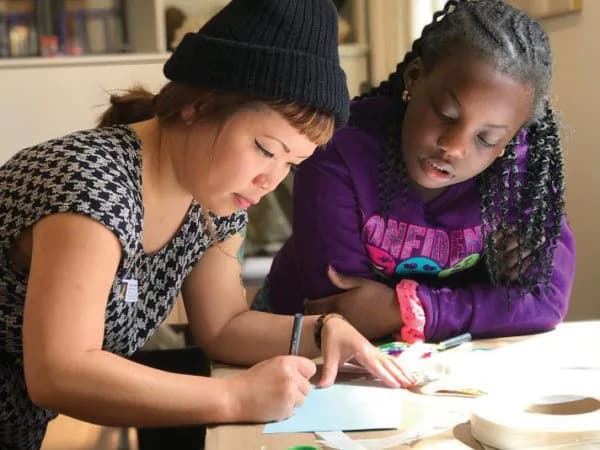 Two young people wearing winter hats and colorful clothing lean over a table, focused on an activity or project together.