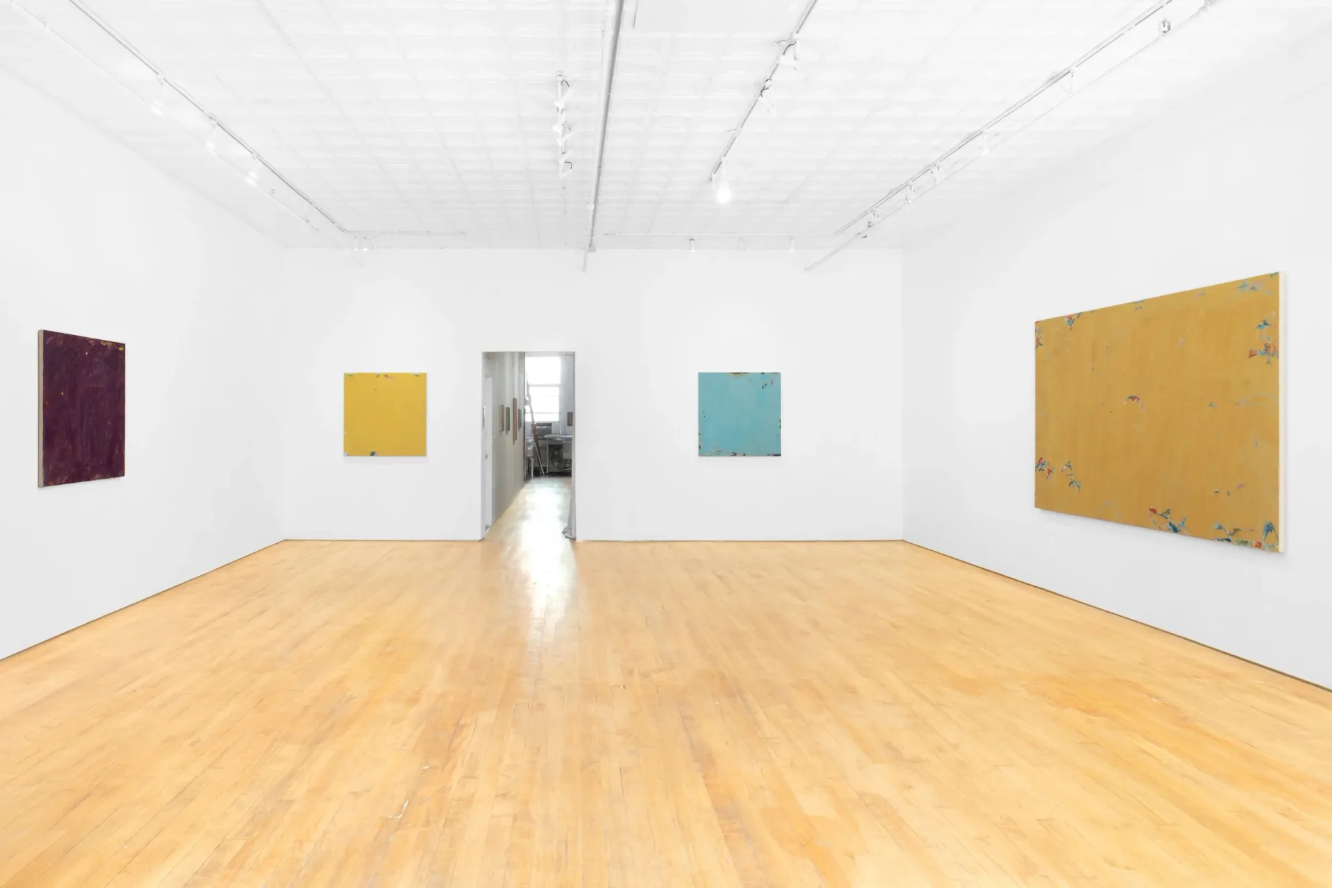 Minimalist gallery room with wooden floor, white walls, and five small to medium-sized colored panel paintings hung sparsely.