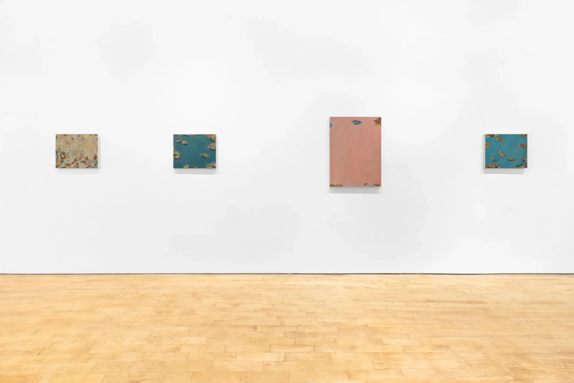 Minimalist gallery wall with wooden floor showing four small framed pieces in muted tones—beige, teal, coral, and blue-green—arranged horizontally.