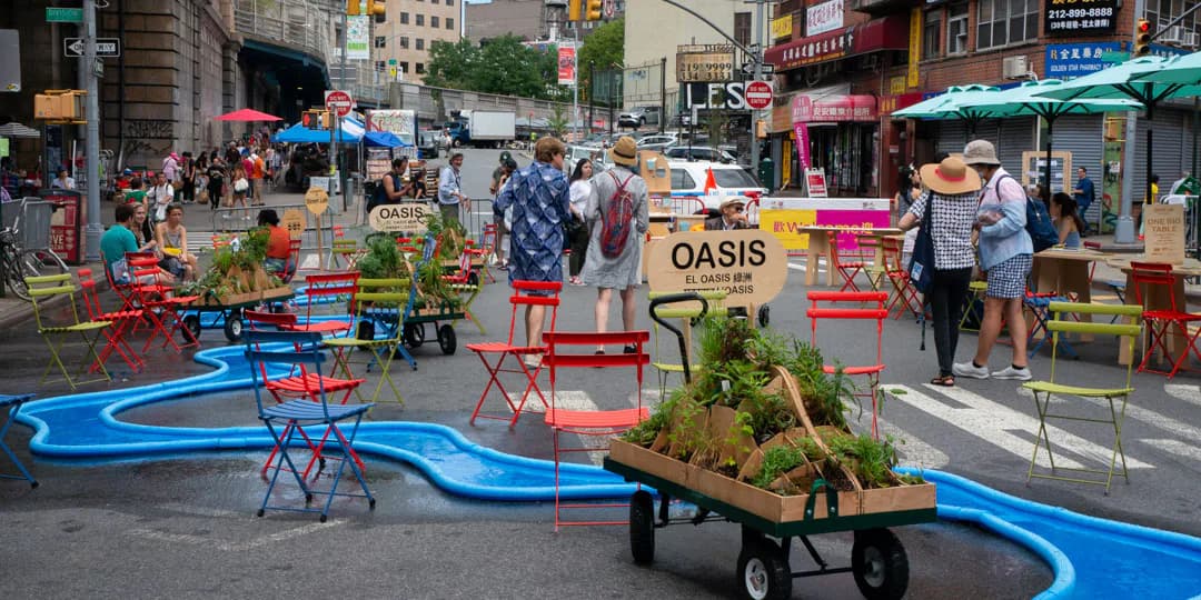 Urban street plaza with colorful painted ground markings, benches, and a wooden wagon labeled 'OASIS' surrounded by pedestrians and buildings.
