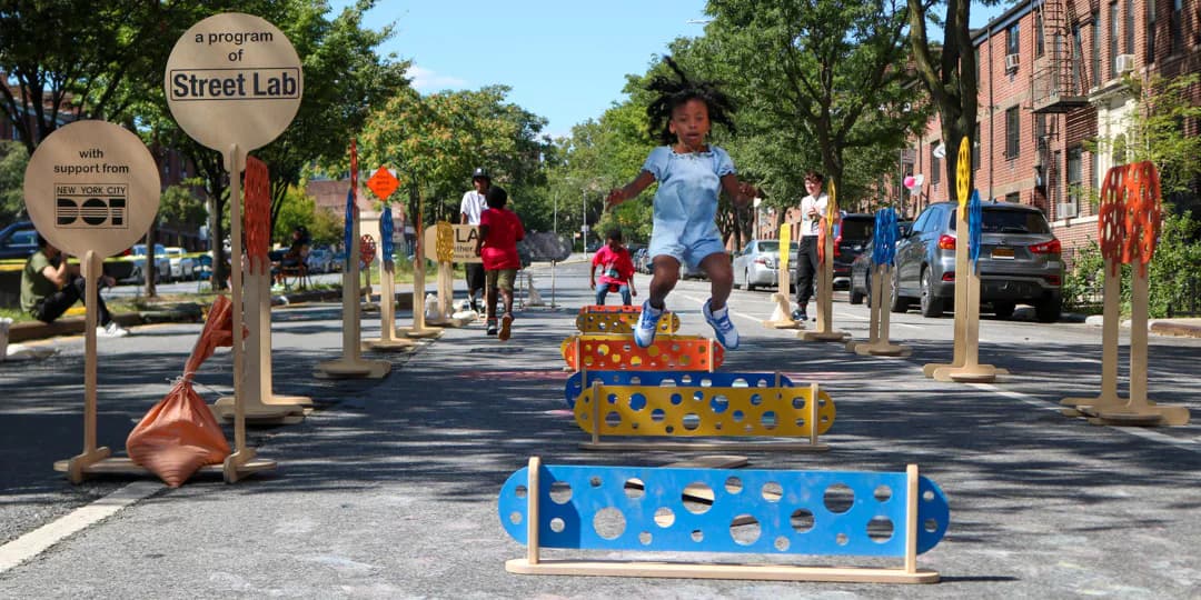 Street Lab mixed-media installation featuring colorful interactive game-like structures and seating elements arranged on a tree-lined urban street with visitors.