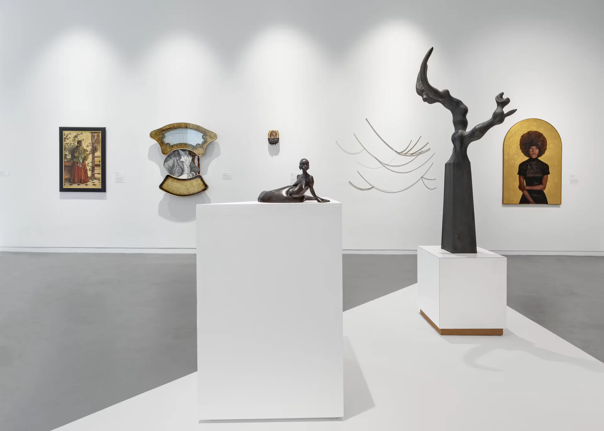 Gallery interior with white walls and gray platforms displaying sculptures, paintings, and mixed-media artworks under gallery lighting.