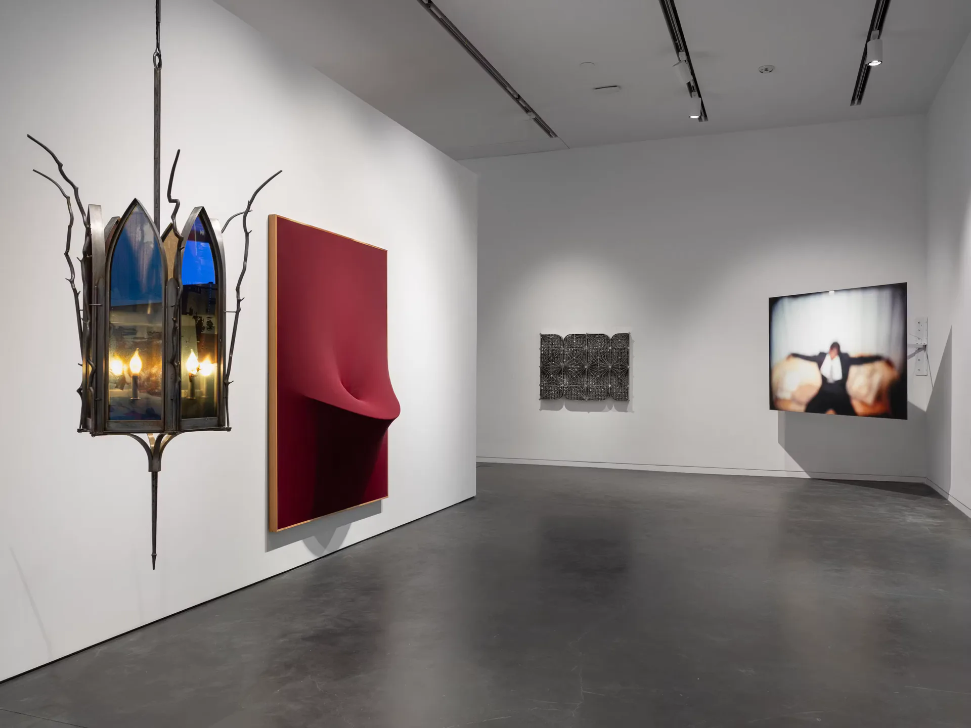 Gallery interior with polished concrete floor showing wall-mounted artworks including a red sculptural piece, lantern installation, and framed photographs.