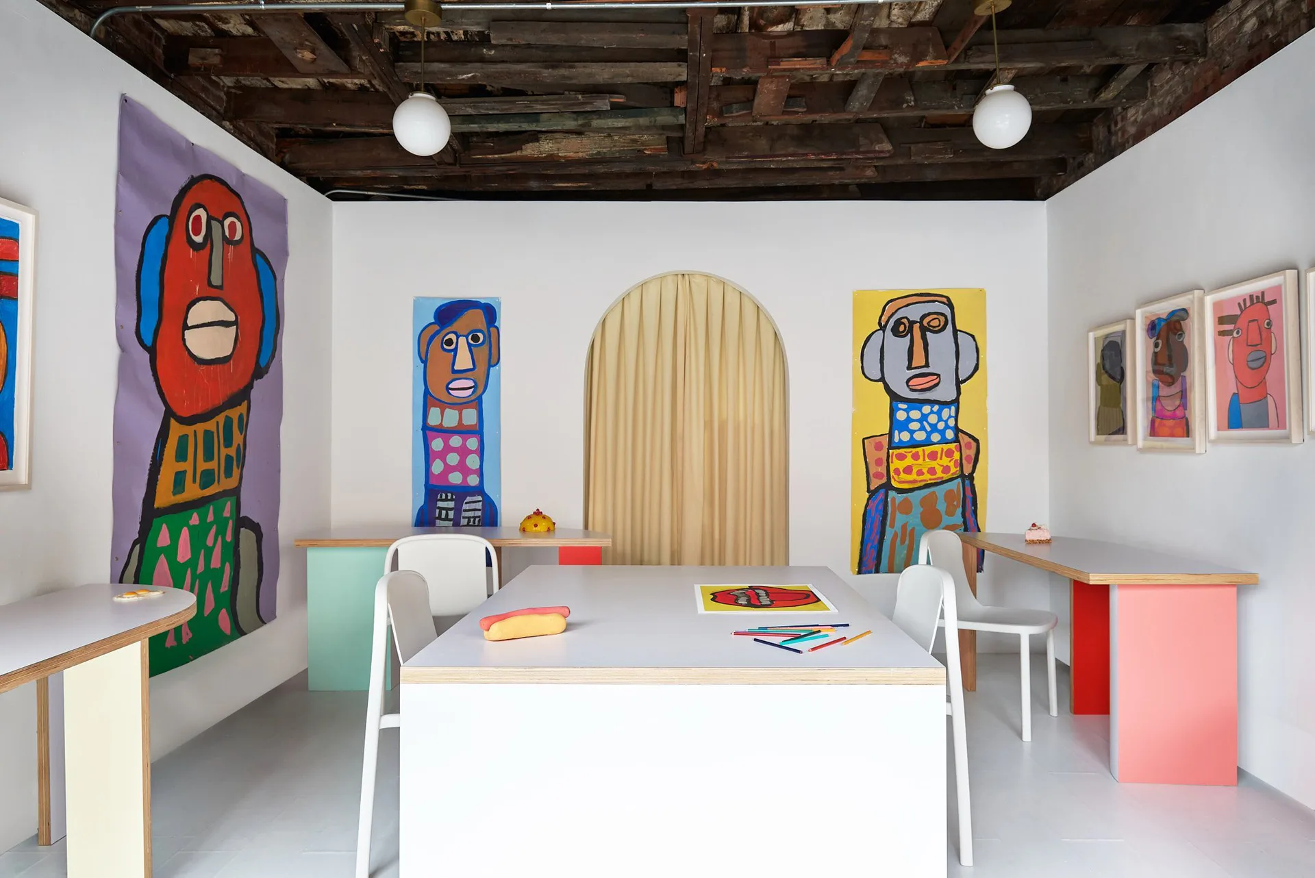 Summertime Gallery interior with white walls, exposed wooden ceiling, colorful figurative paintings, a white table with chairs, and a natural wood arched doorway.