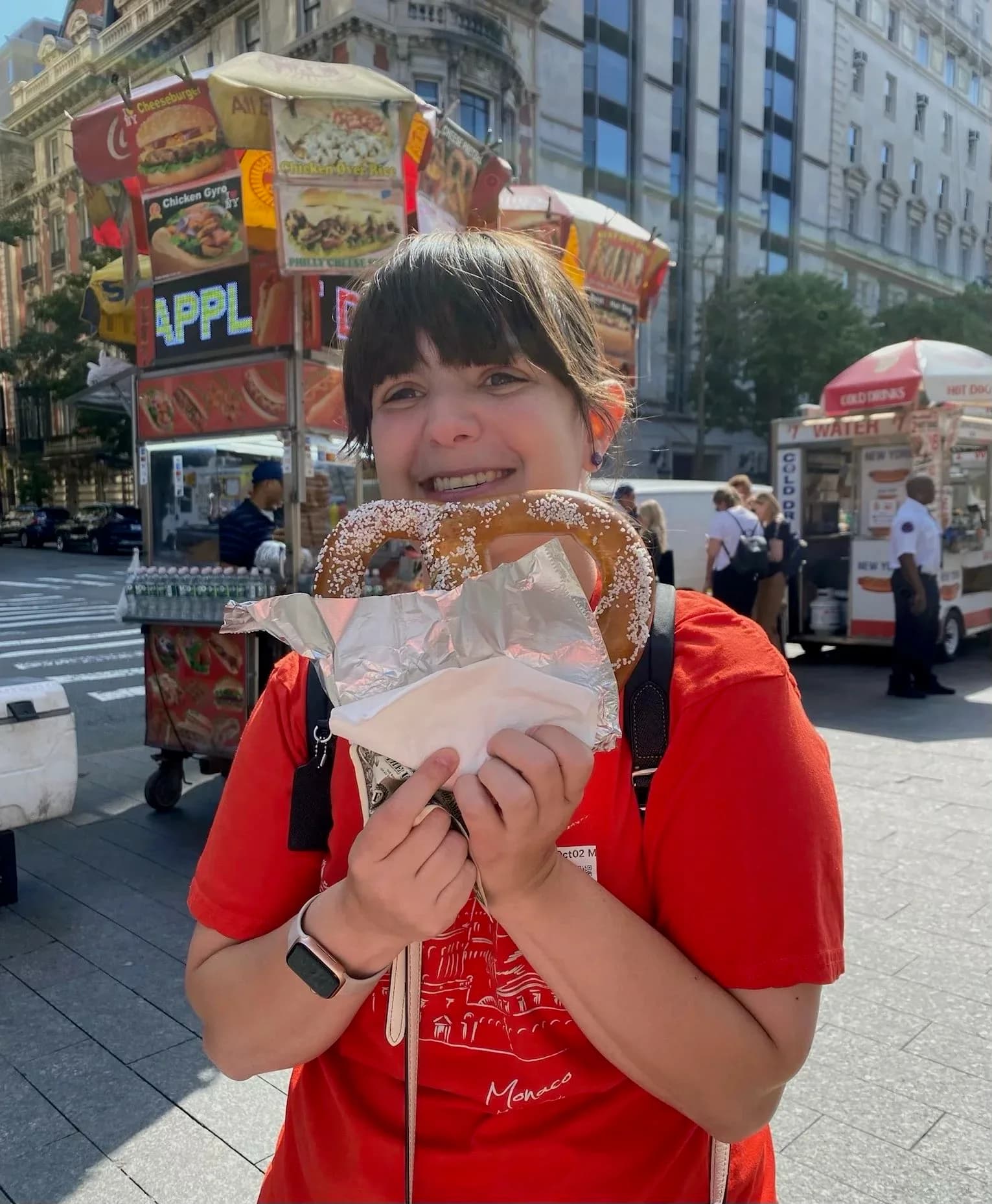 Child holding a large pretzel in an urban public square with food vendor signage and buildings visible in the background.