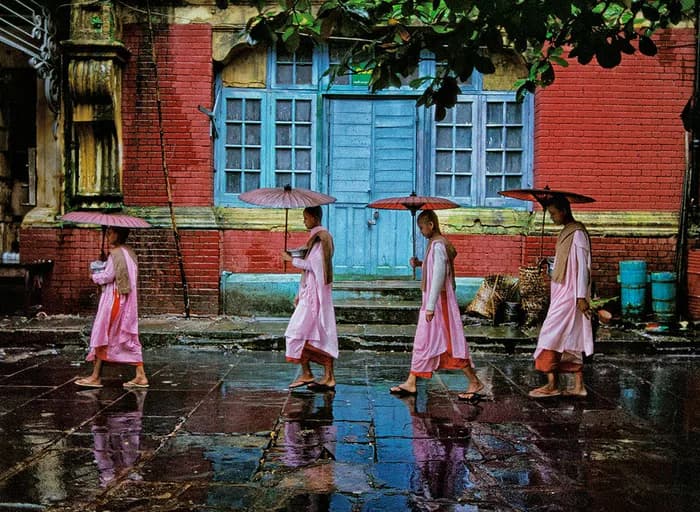 Four figures in pink dresses walking across wet ground reflecting red brick building with blue door and windows.