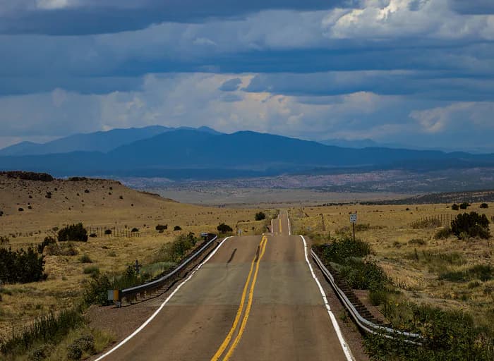 Perspective photograph of straight desert highway with yellow center line leading toward distant mountains under dramatic cloudy sky.