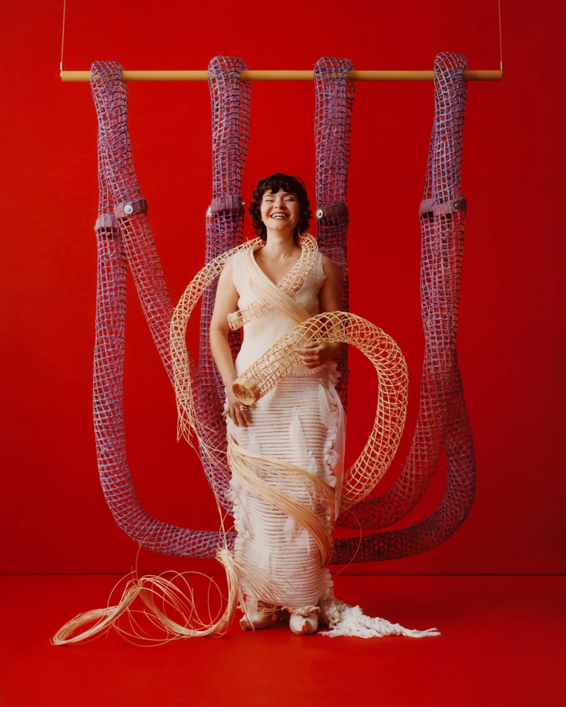 Large-scale mixed-media installation: woman in cream dress poses within geometric rope and metal framework sculpture against vibrant red background.