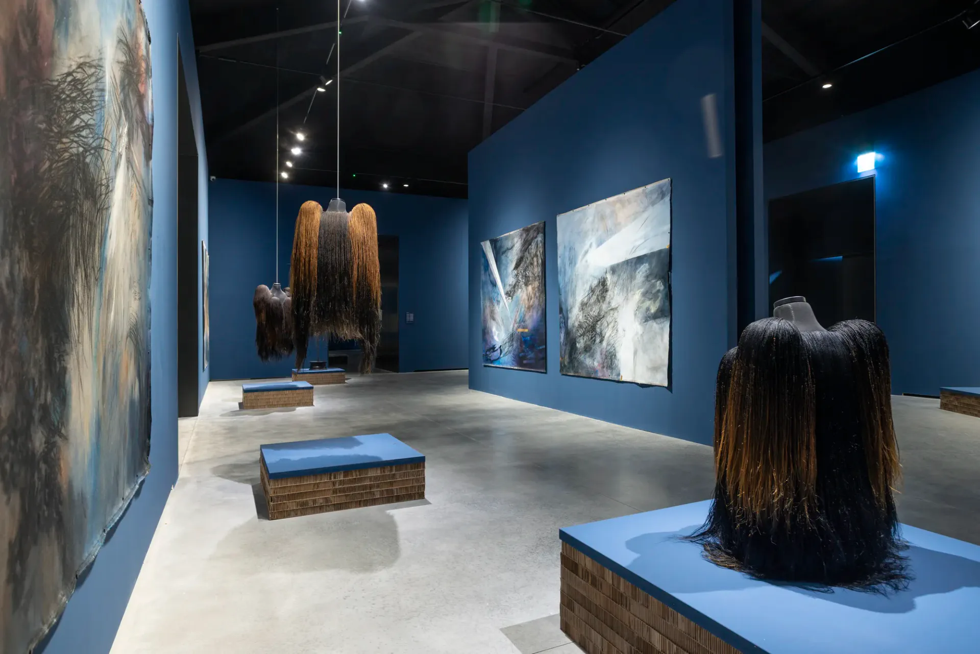 Gallery interior with deep blue walls displaying large-scale photographs and sculptures with long dark hair on blue pedestals.