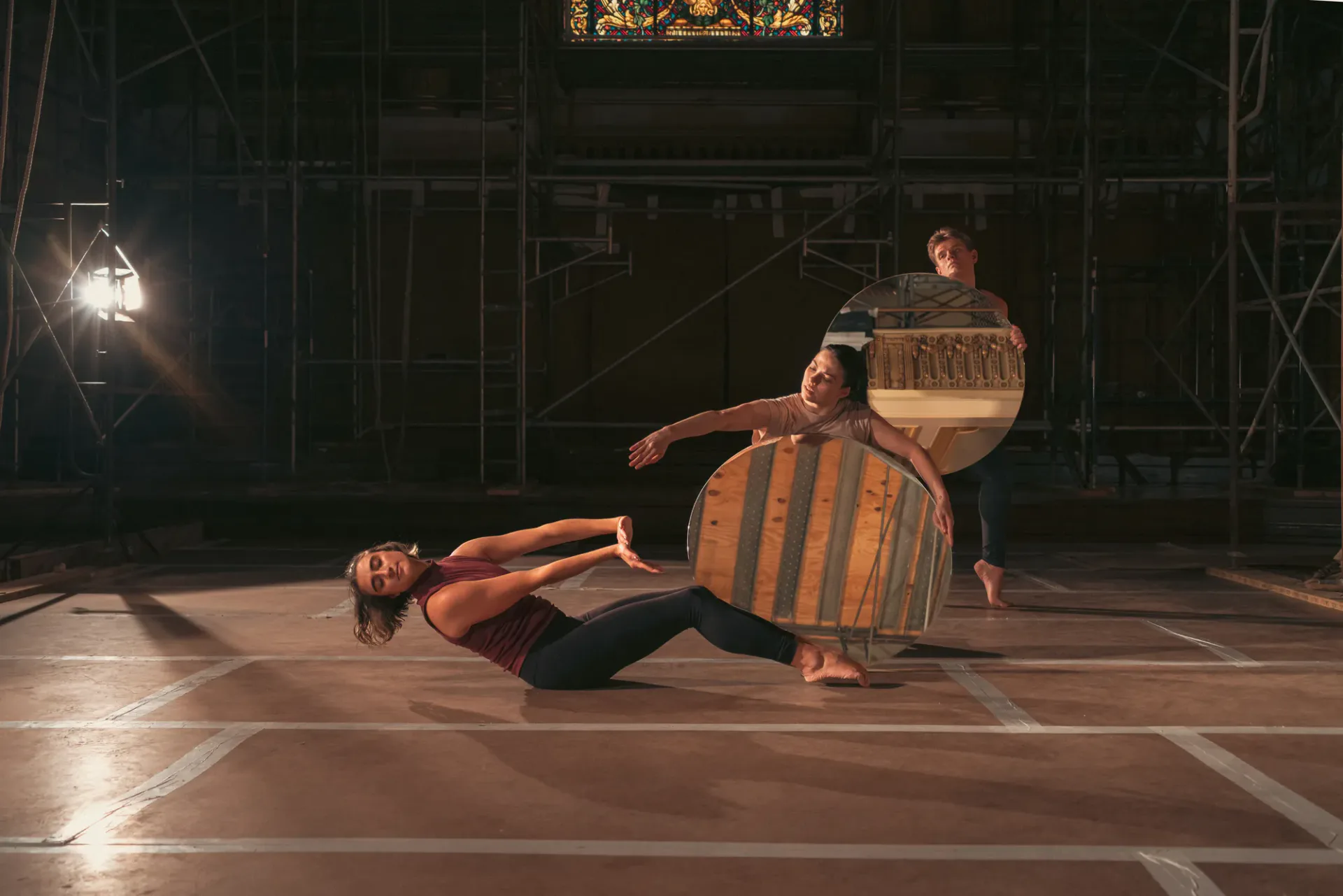 Performance or installation artwork showing two figures in striped clothing positioned on floor with chair and dramatic stage lighting against dark backdrop.