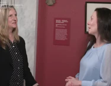 Two women conversing in a gallery interior with a burgundy accent wall and artwork display visible at Swann Auction Galleries.