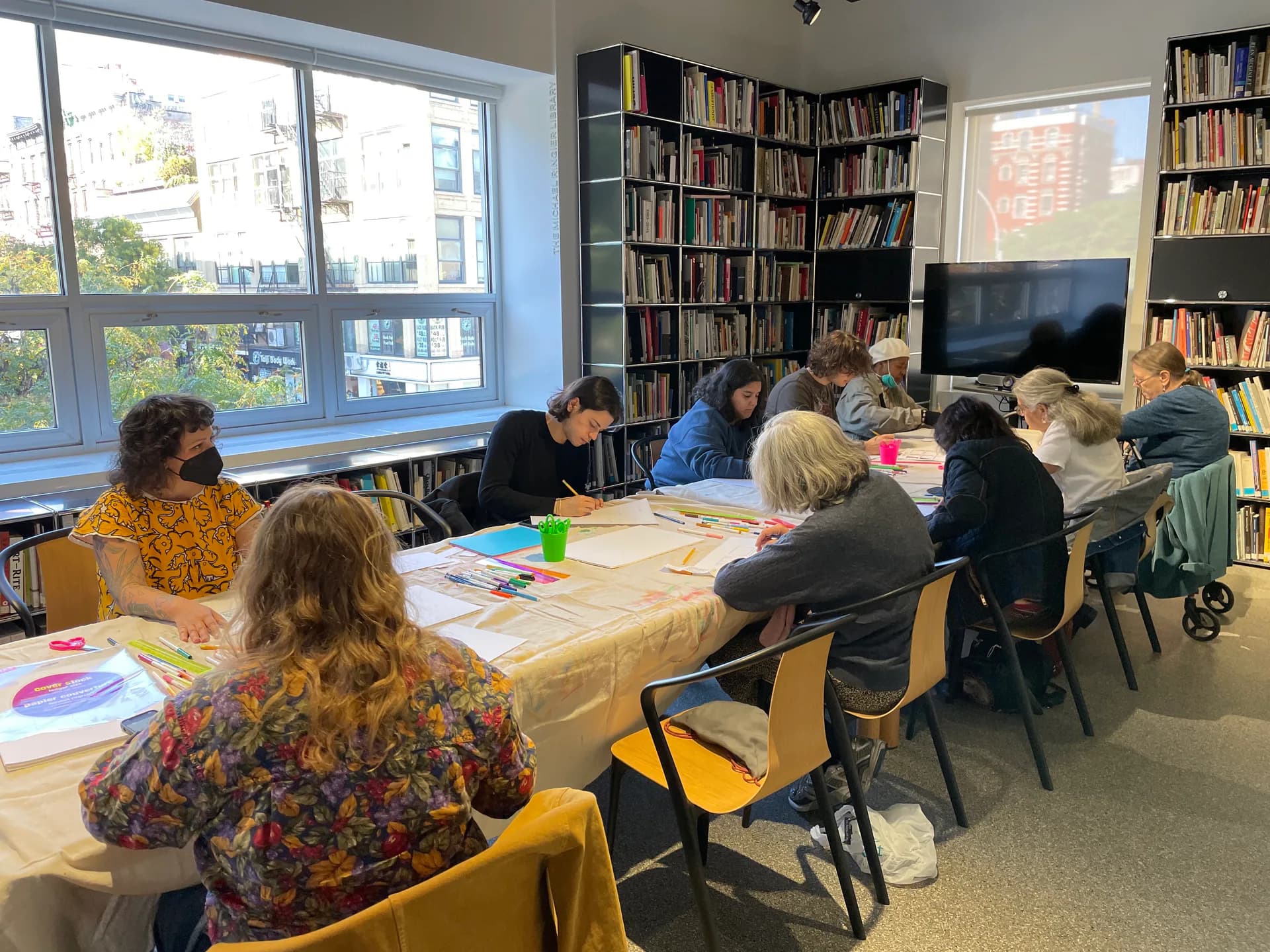 Library or education space with bookshelves lining walls, large windows, and a group seated at a long table with art materials and worksheets.
