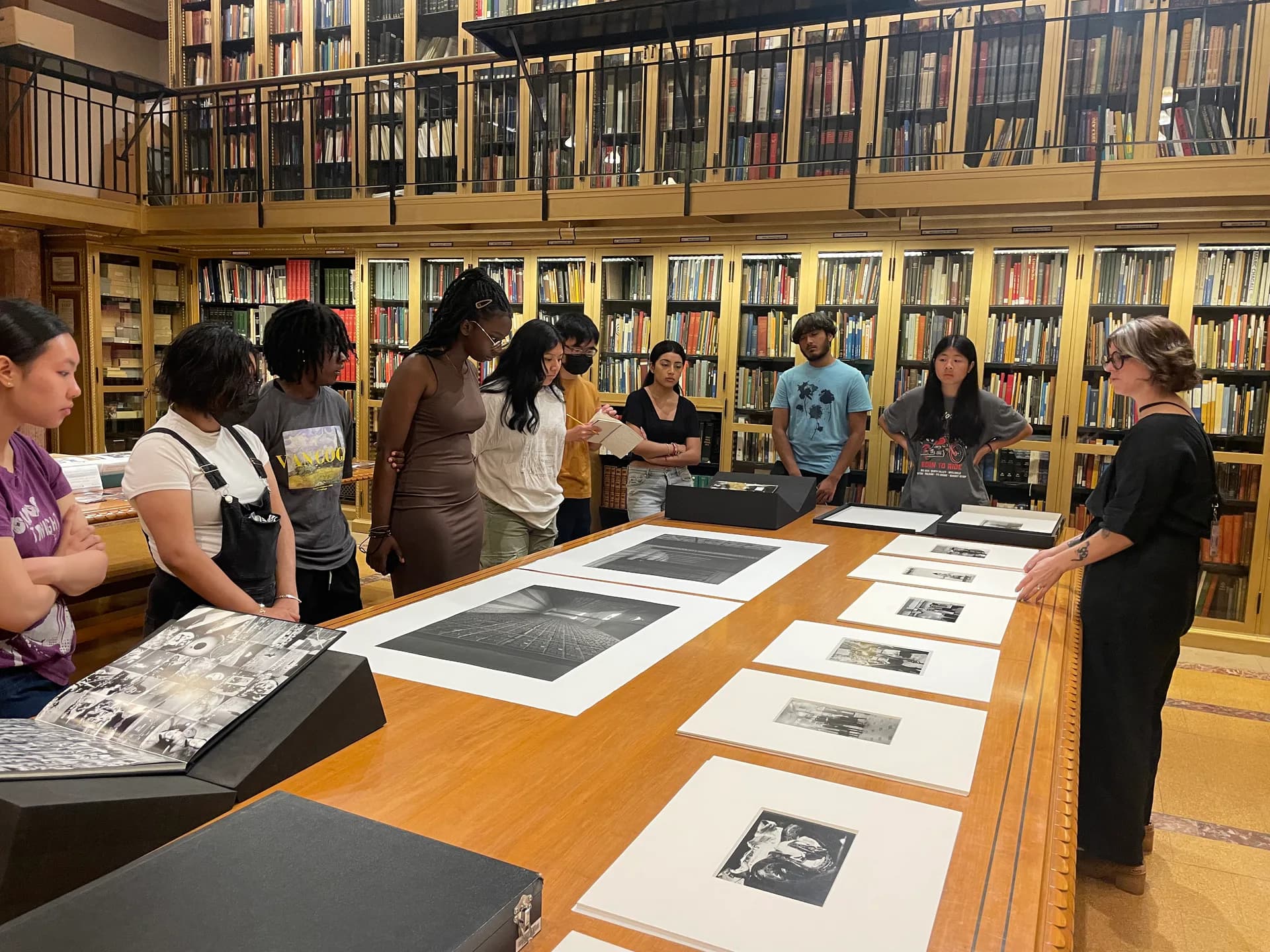 Swiss Institute library with floor-to-ceiling shelves and gallery lighting; visitors viewing black and white photographs and prints displayed on wooden table.