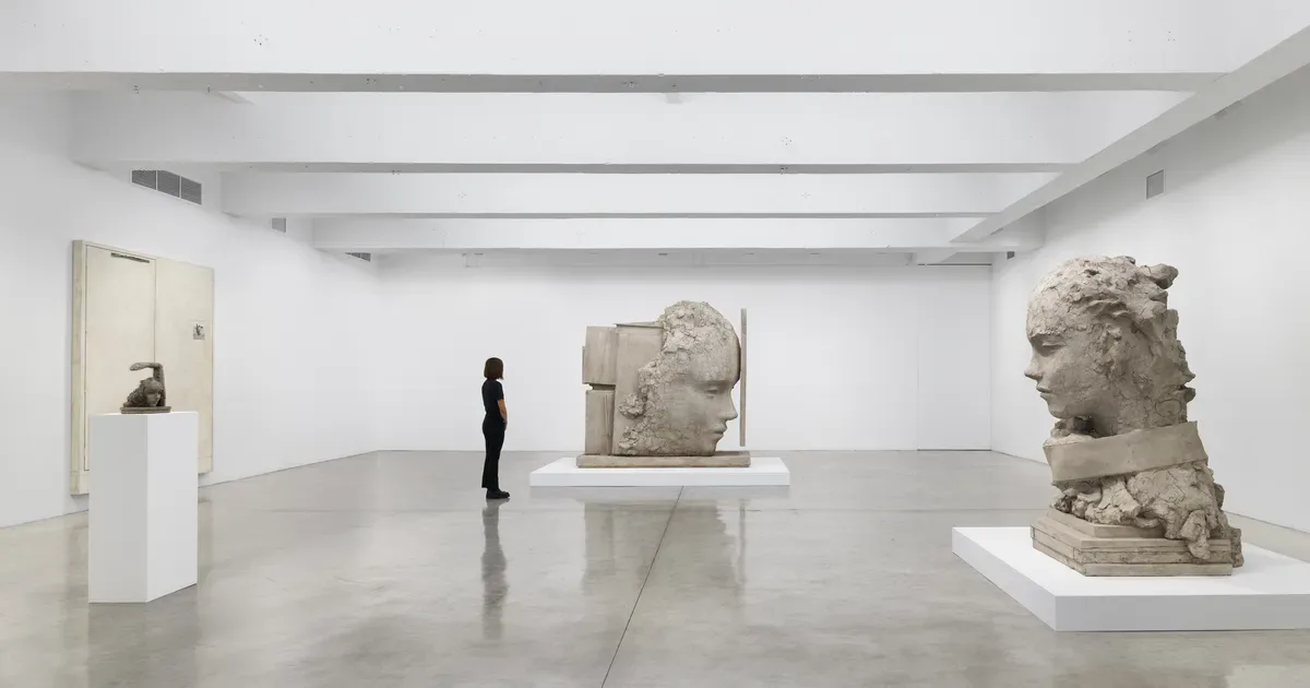 Contemporary art gallery showing three stone sculptural busts positioned on illuminated white platforms in a sparse, minimalist setting.
