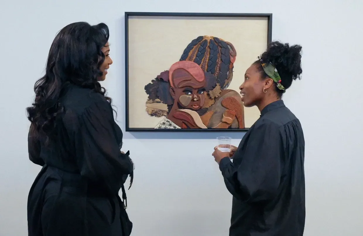 Two people viewing a framed painting of a figure wearing a hat and orange garment on a white gallery wall.