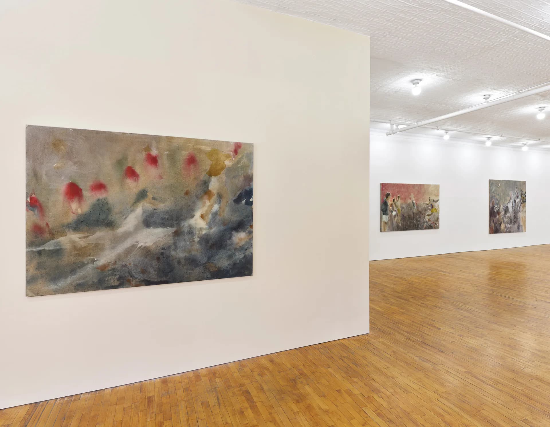 Gallery interior with white walls and wooden floor, displaying three framed mixed-media artworks at varying heights showing abstracted figures and atmospheric elements.