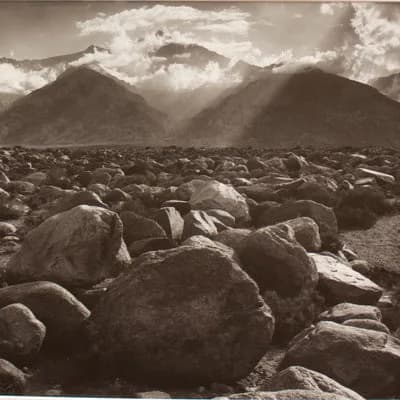 Black and white landscape photograph of rocky terrain with dramatic mountain peaks and clouded sky in the distance.
