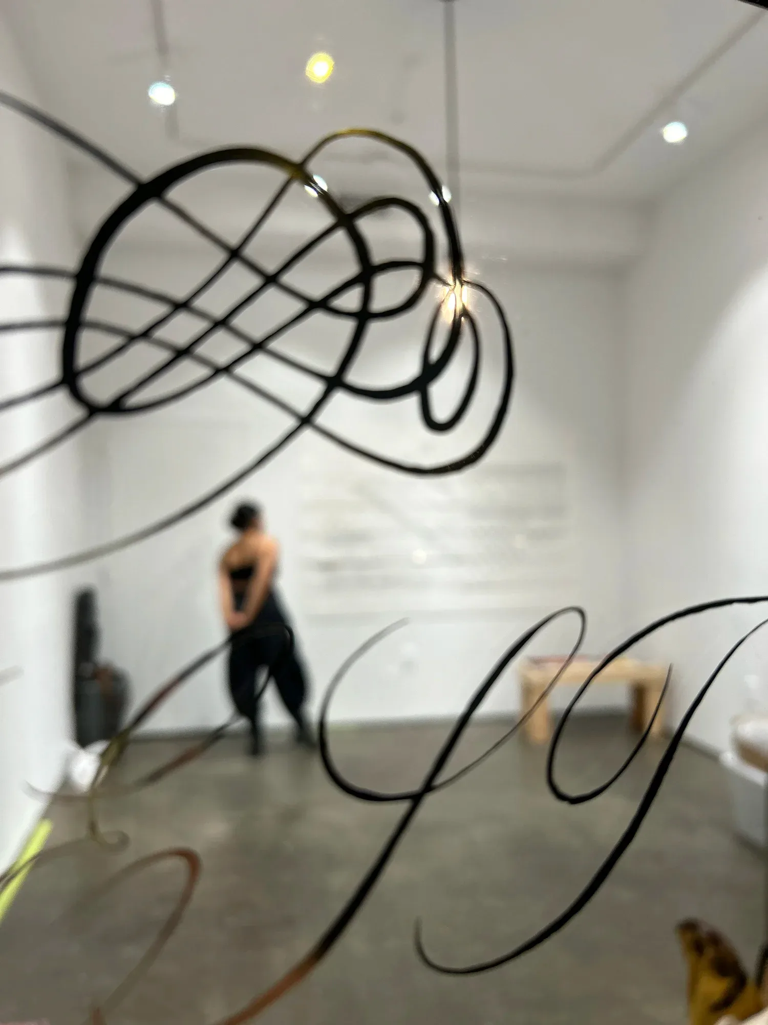 Black-line kinetic wire sculpture suspended in white gallery space, forming abstract flowing loops; figure in background.