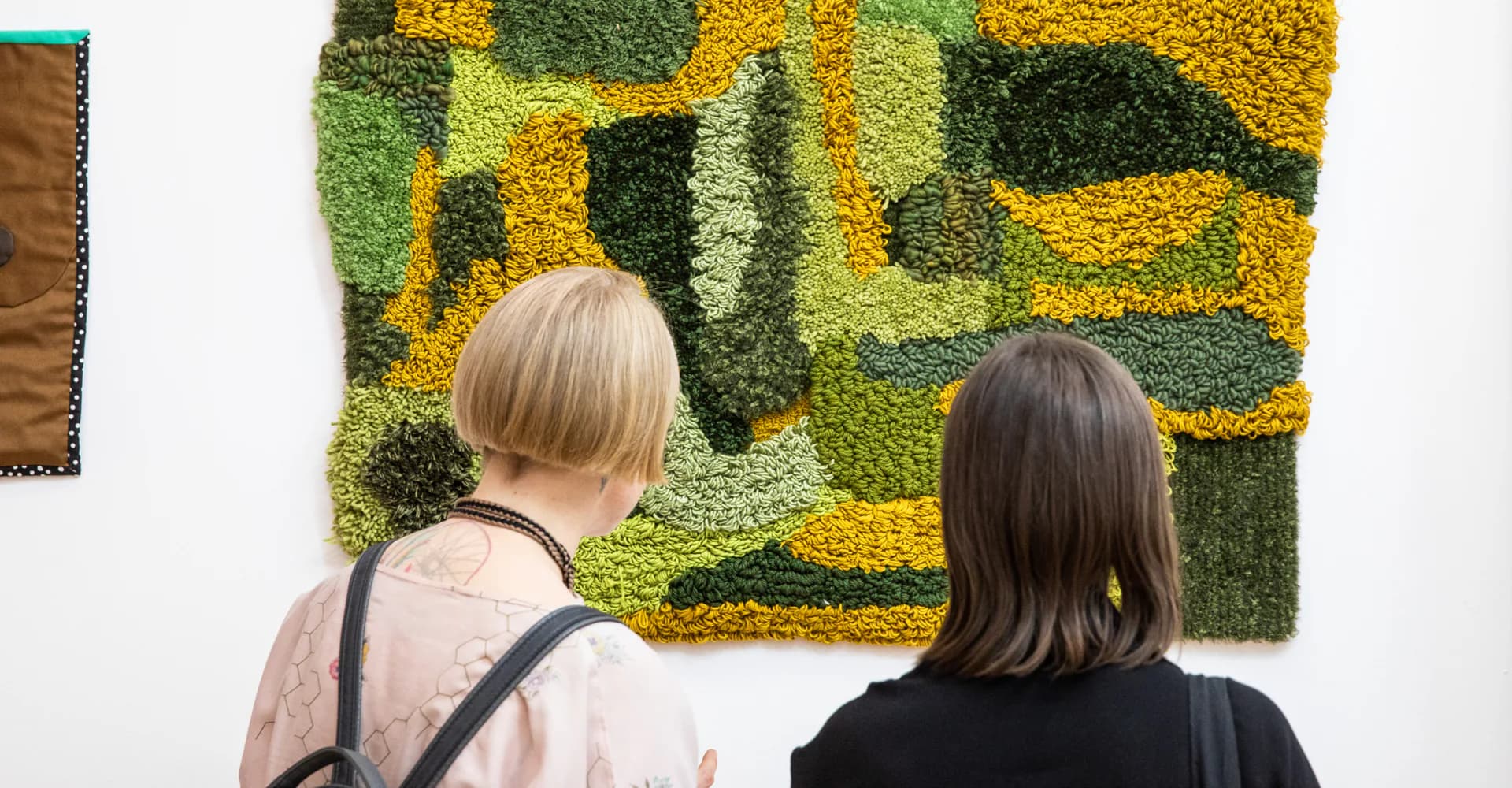 Two visitors viewing a vibrant yellow and green abstract fiber artwork mounted on white gallery wall.