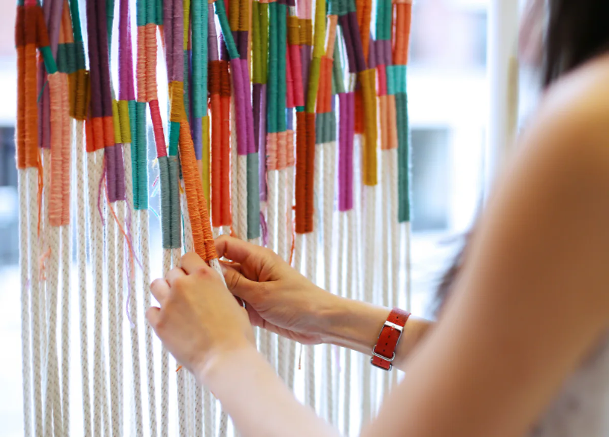 Person's hands selecting colorful yarn strands from a hanging display of dyed textile fibers.