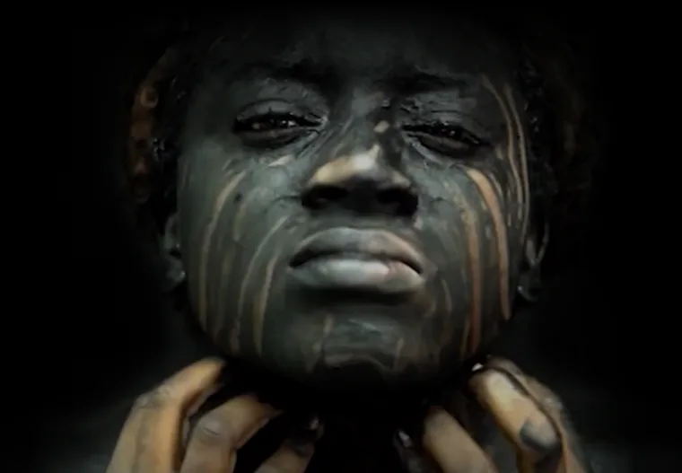 Sculptural portrait head in dark patinated bronze or similar metal, with vertical striations across the face and a contemplative expression, against black background.