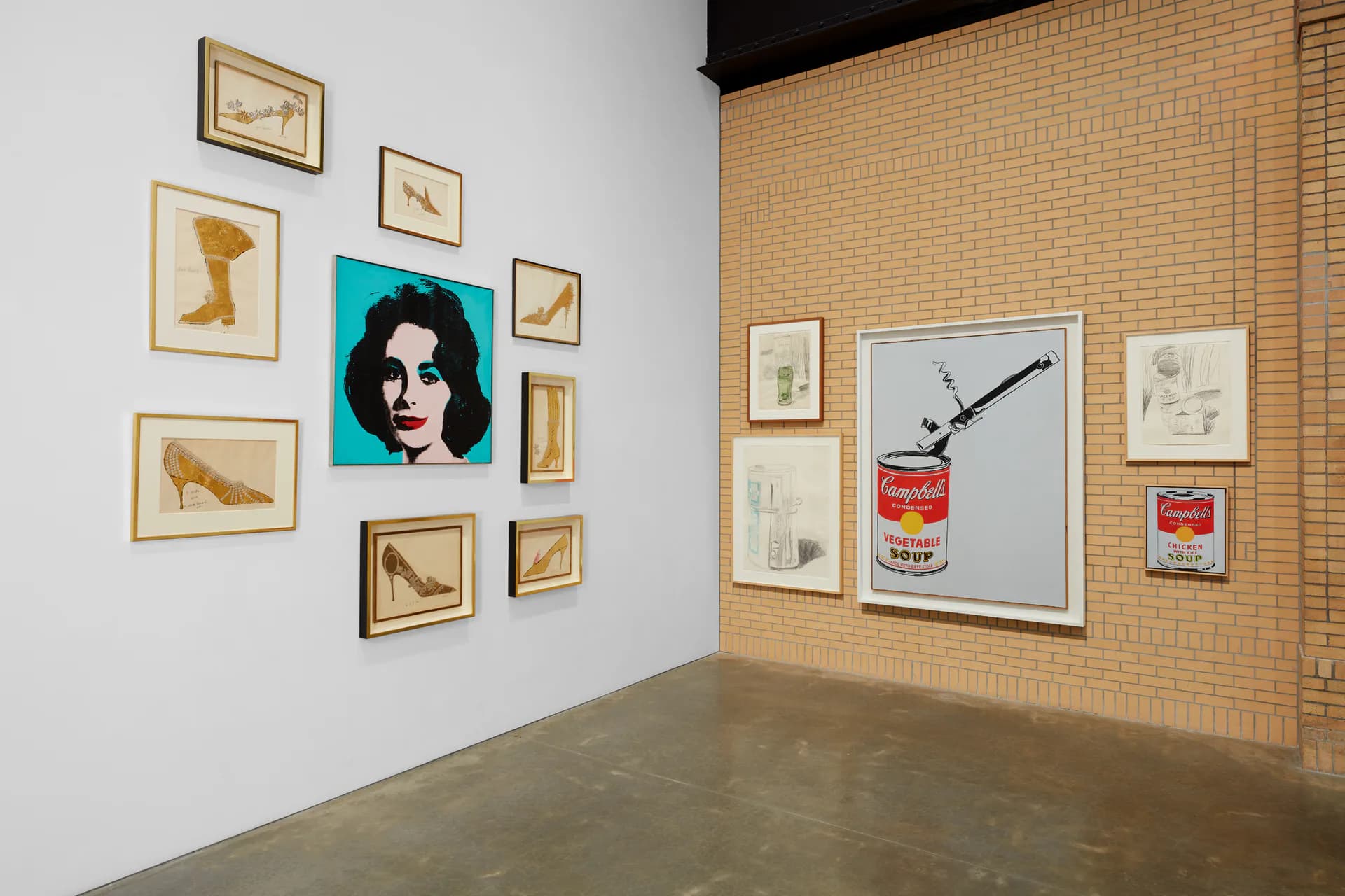 Gallery corner showing white and brick walls with a grid arrangement of framed drawings on the left and a larger pop art portrait with soup cans imagery on the brick wall to the right.