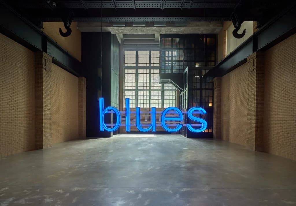 Neon blue cursive text reading 'blues' installed in a gallery space with brick walls, concrete floor, and industrial windows visible in background.