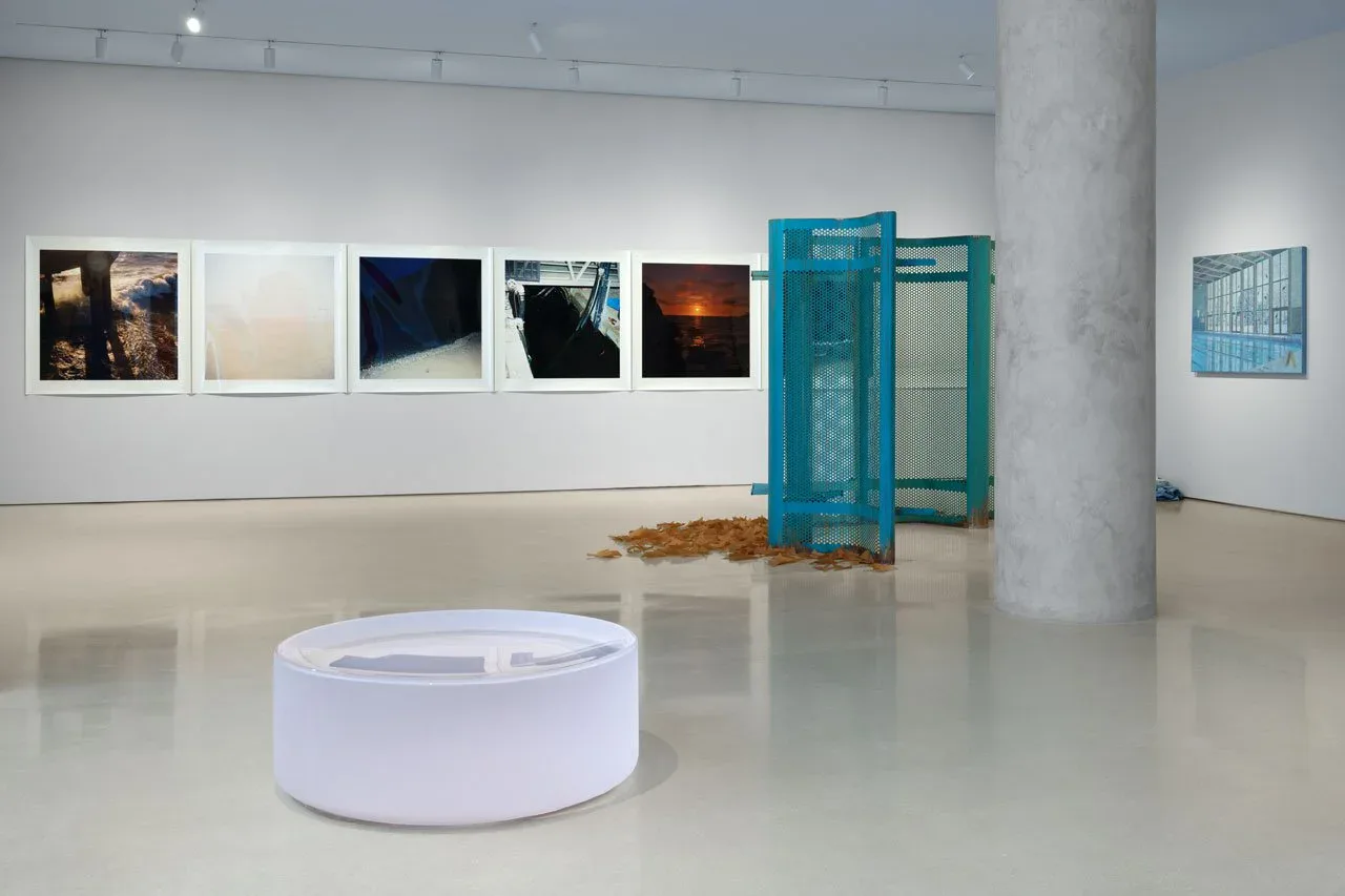 Contemporary gallery interior with white walls, polished concrete floor, displaying framed artworks in a row and a turquoise mesh sculptural installation.