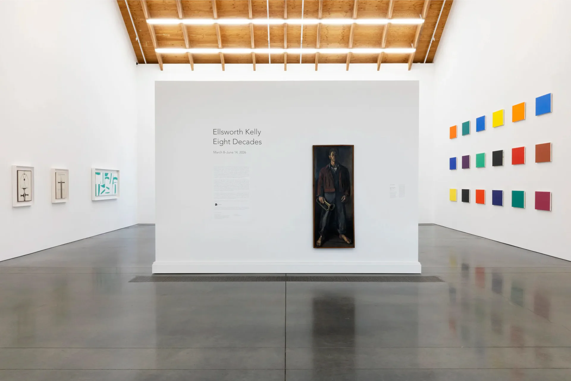 Gallery space with wooden ceiling grid, central white pedestal with black artwork, color swatches on right wall, and small works on left.