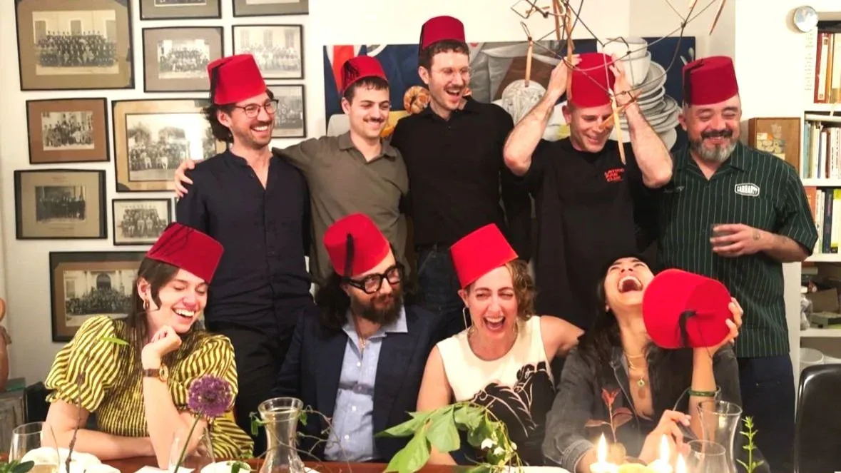 Group photo of nine people wearing matching red fez hats indoors, some standing and some seated, in front of shelves with framed photographs.