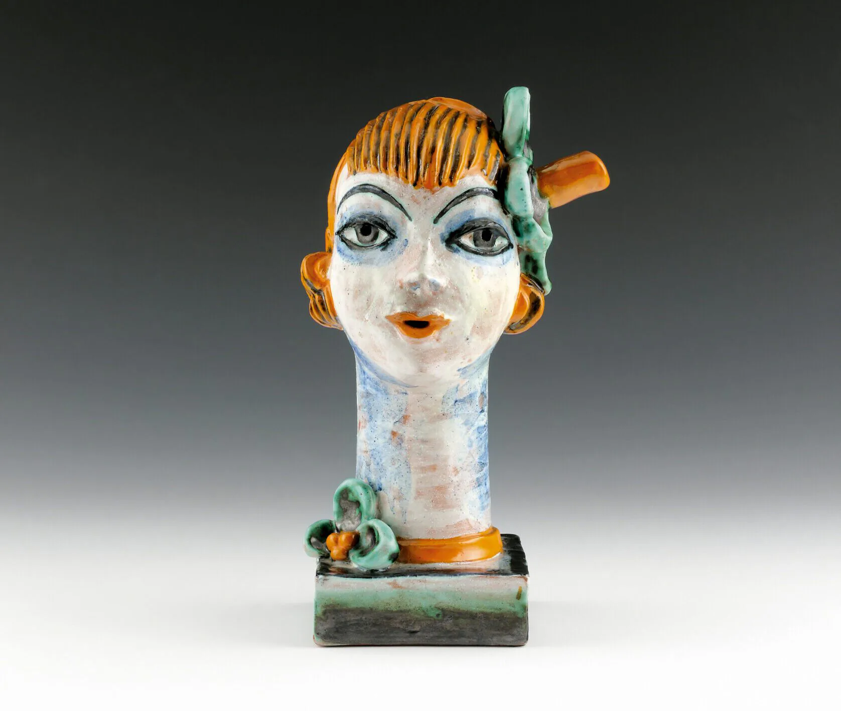 Ceramic sculptural bust featuring a stylized female head with orange hair, white face, blue eyes, and orange and green decorative elements on a base.