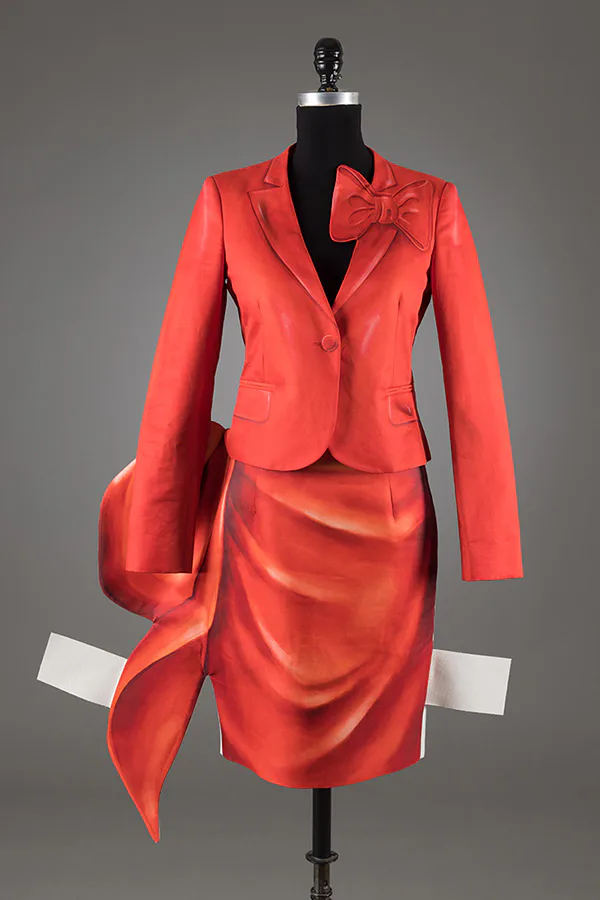 Bright coral-red silk dress suit on dress form, featuring a cropped blazer with bow detail and draped skirt with flowing panel.