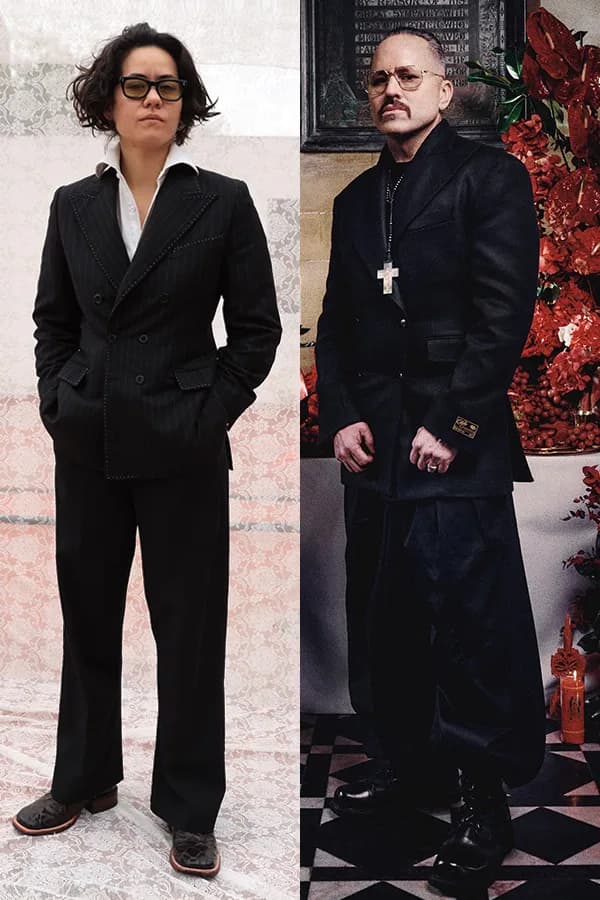 Two people in black double-breasted blazers; left figure wears glasses, right figure wears cross necklace and glasses.